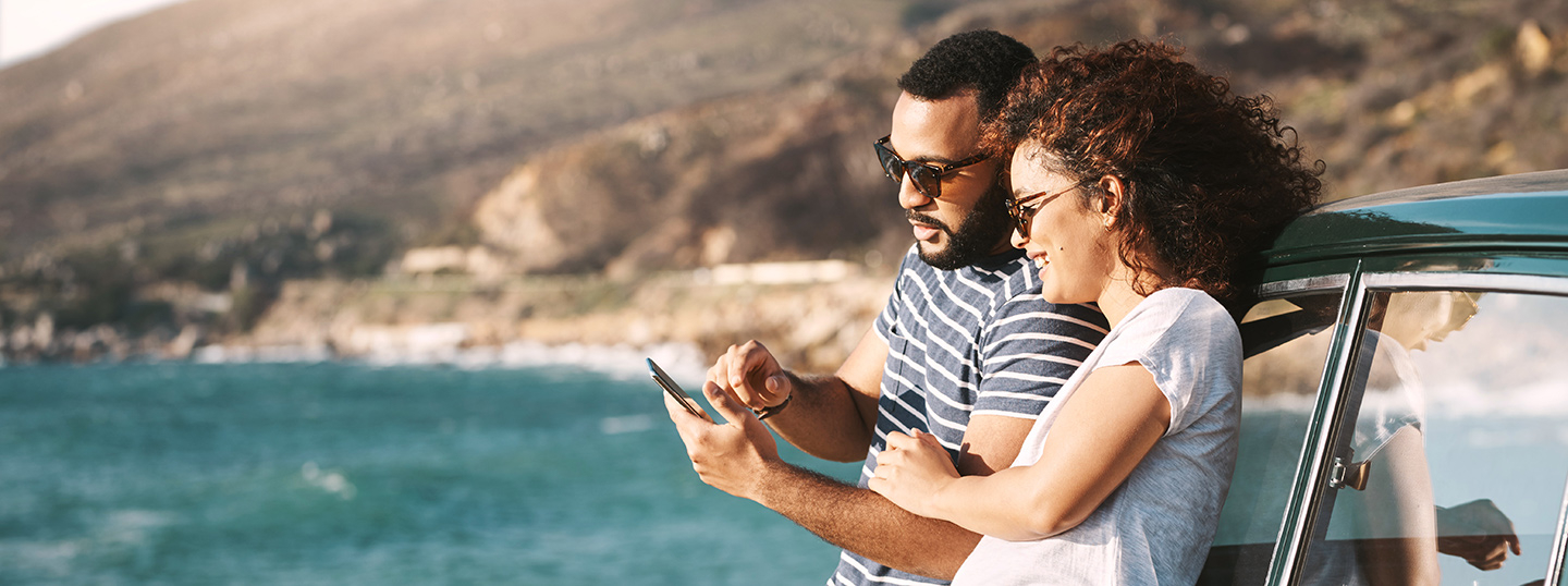 Couple leaning agains their car by the coast, looking at a smartphone