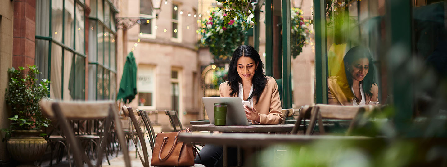 Woman sat outside a coffee shop in a modern, public area, confidently looking at her laptop